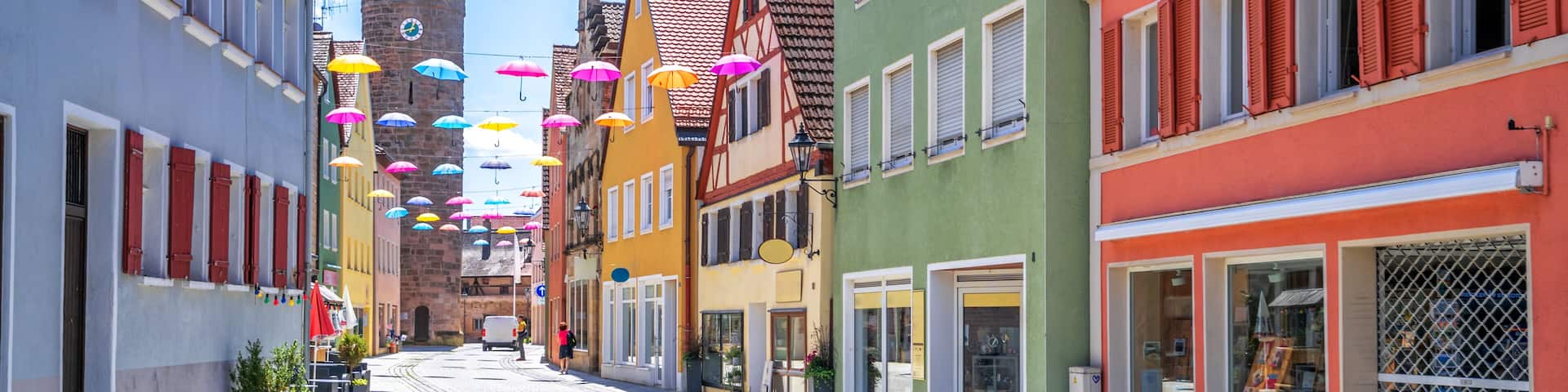 A cobblestone street lined with colorful buildings, a tower, and string lights with umbrellas.