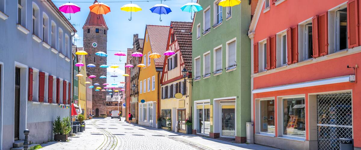 A cobblestone street lined with colorful buildings, a tower, and string lights with umbrellas.