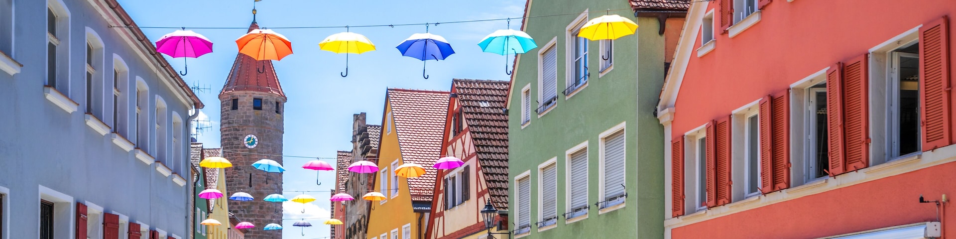 A cobblestone street lined with colorful buildings, a tower, and string lights with umbrellas.