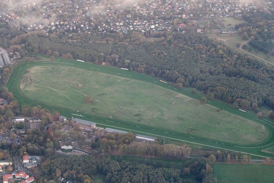 Eine Vogelperspektive auf ein großes, grünes Sportfeld mit einer Laufbahn, umgeben von dichten Wäldern und einer kleinen Stadt mit Gebäuden und Straßen.