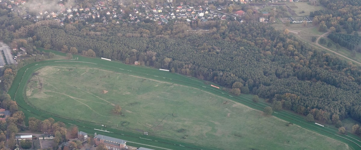 An aerial view of a large green sports field with a running track, surrounded by dense forests and a small town with buildings and roads.