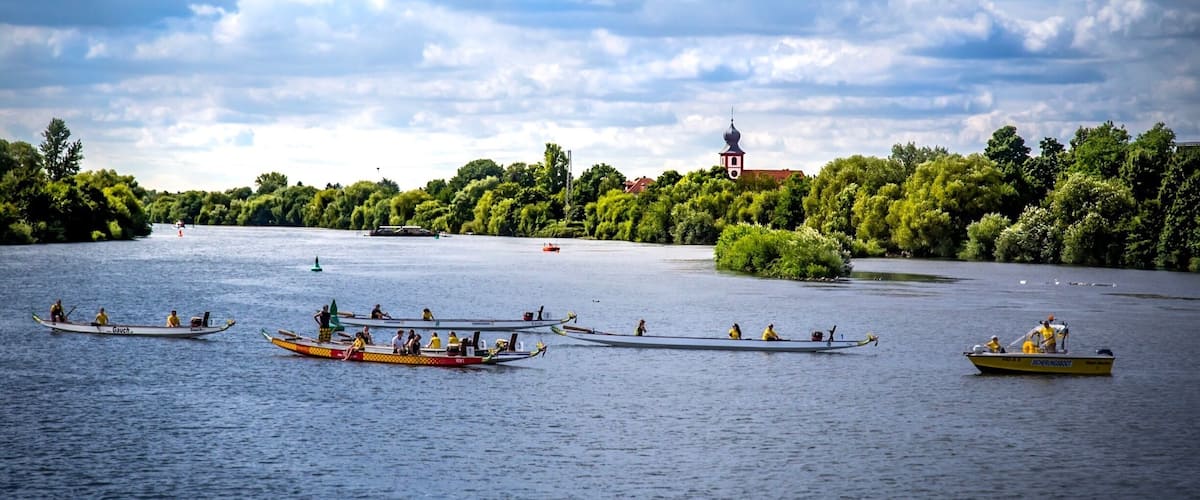 A calm lake with several traditional boats, a distant church, and lush greenery along the shore.