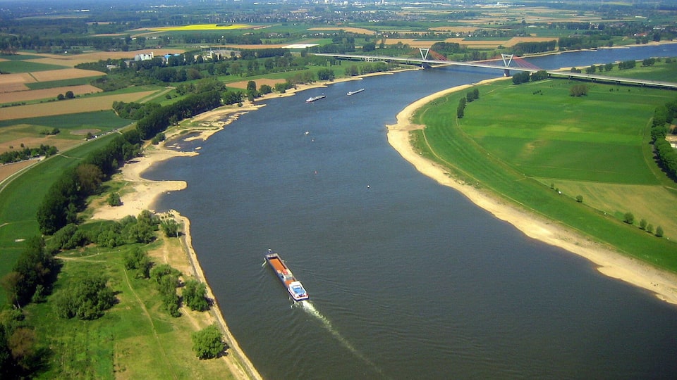 A river with a cargo ship, surrounded by green fields and a bridge in the distance.