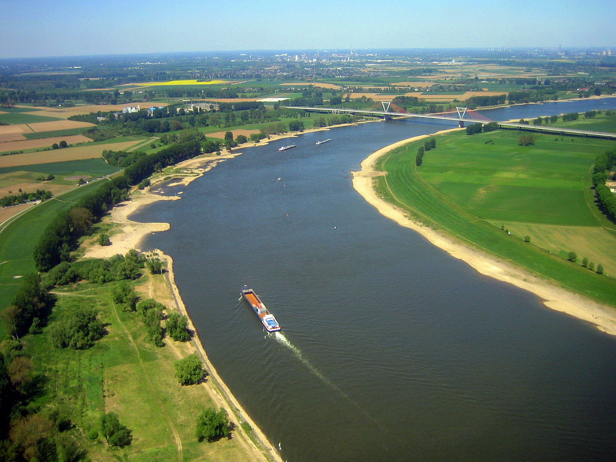 A river with a cargo ship, surrounded by green fields and a bridge in the distance.