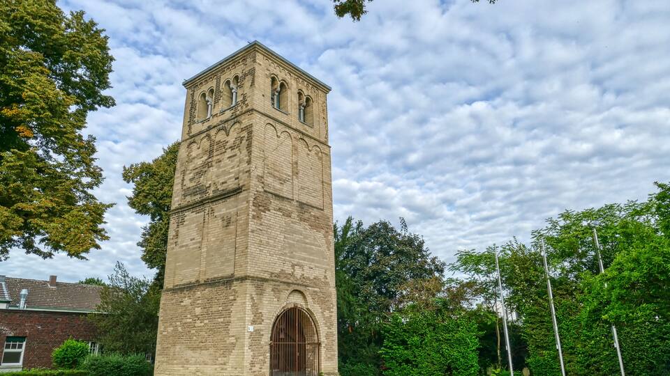A historic stone tower with arched windows and a large door, surrounded by lush greenery and trees.
