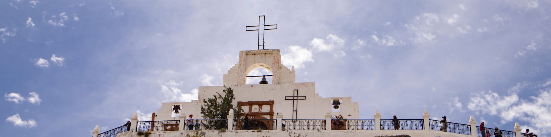 Una iglesia blanca con torre de campanas y cruz sobre una colina rocosa bajo un cielo parcialmente nublado.
