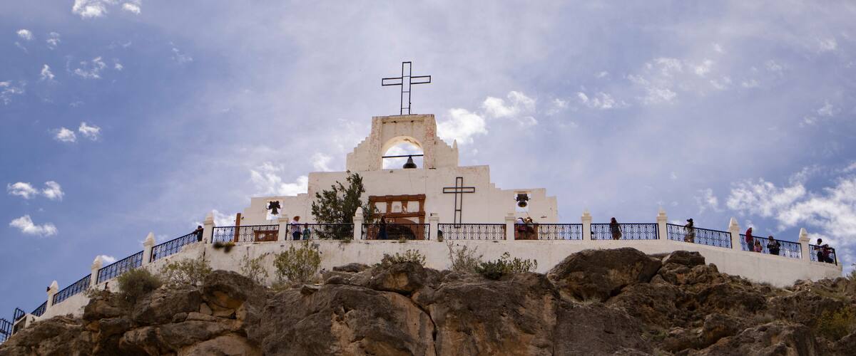A white church with a bell tower and cross on a rocky hill under a partly cloudy sky.