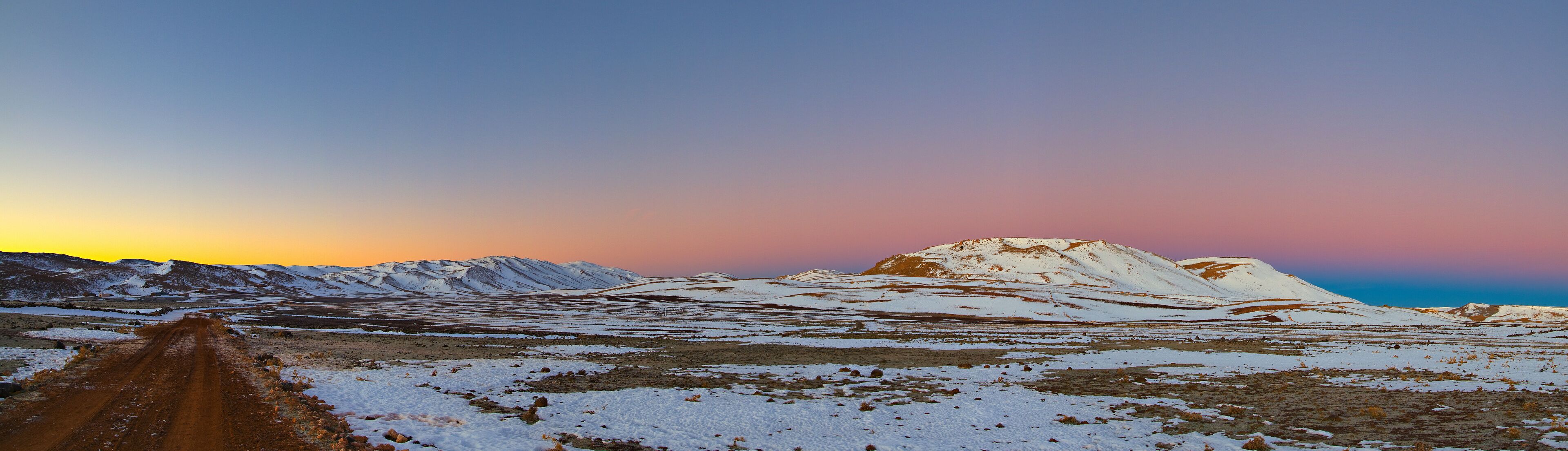 A snow-covered landscape with a dirt road leading towards a mountain range during sunset.