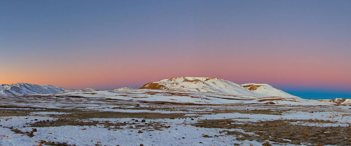 A snow-covered landscape with a dirt road leading towards a mountain range during sunset.