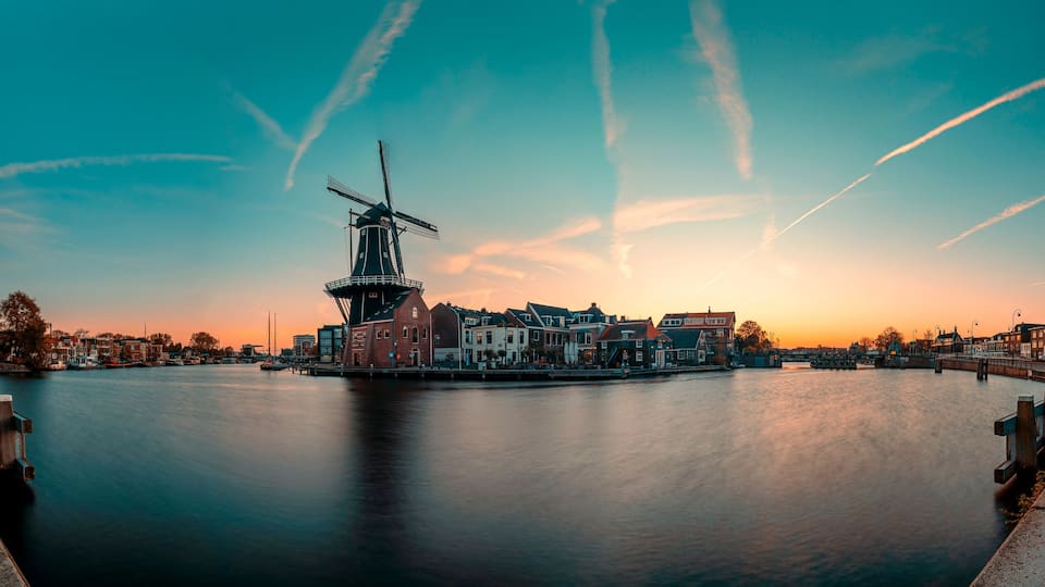 A calm river with a windmill and historic buildings at sunset.