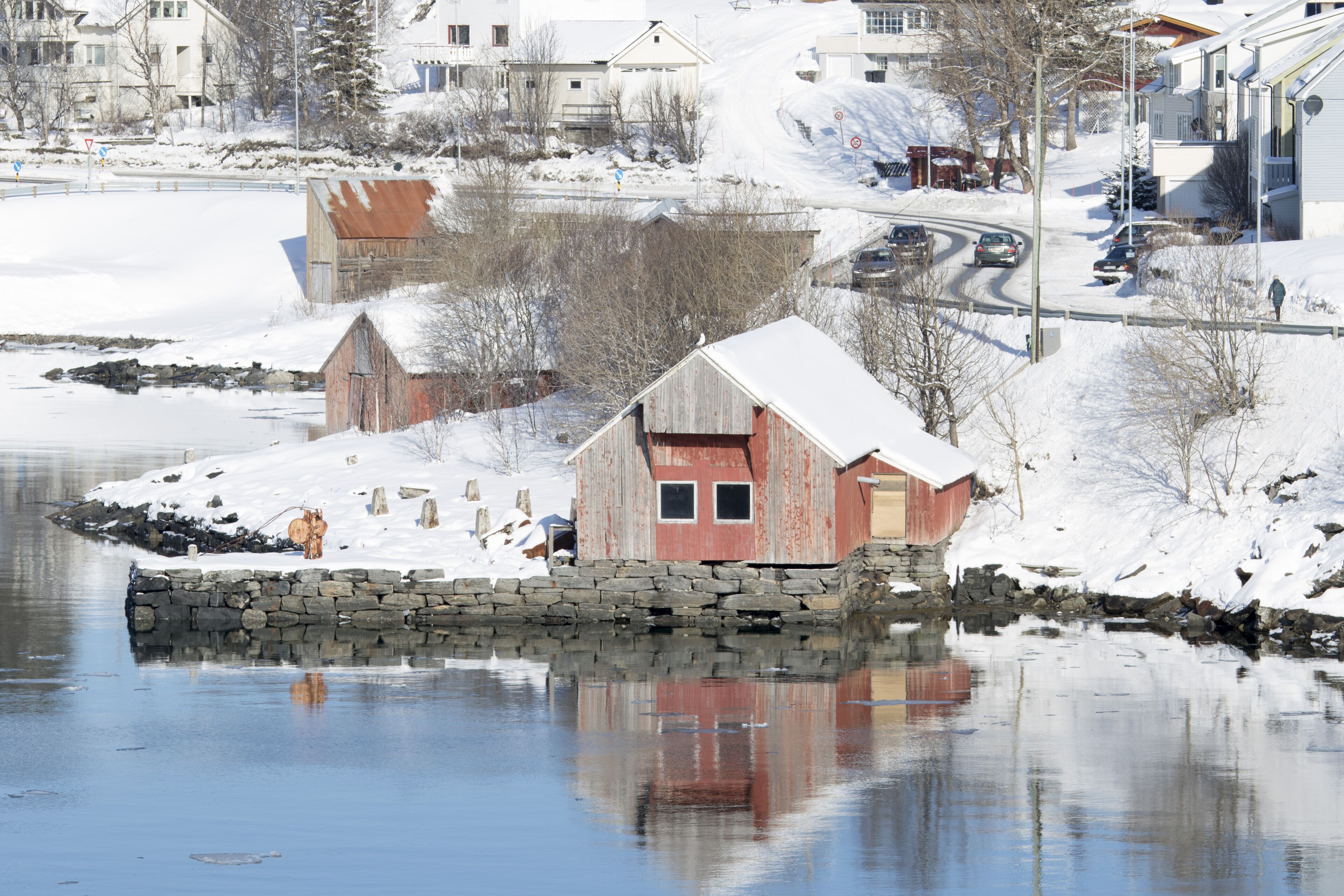 A wooden boathouse by a frozen lake, surrounded by snow-covered houses and trees.