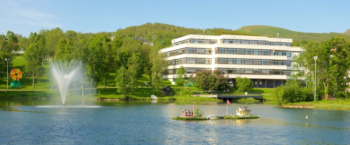 A lake with a fountain, a boat, and a modern building surrounded by greenery and hills.