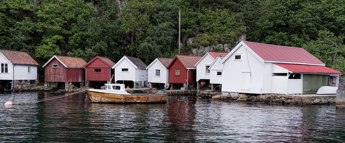 A row of wooden houses with red roofs by a calm body of water, surrounded by dense forest.