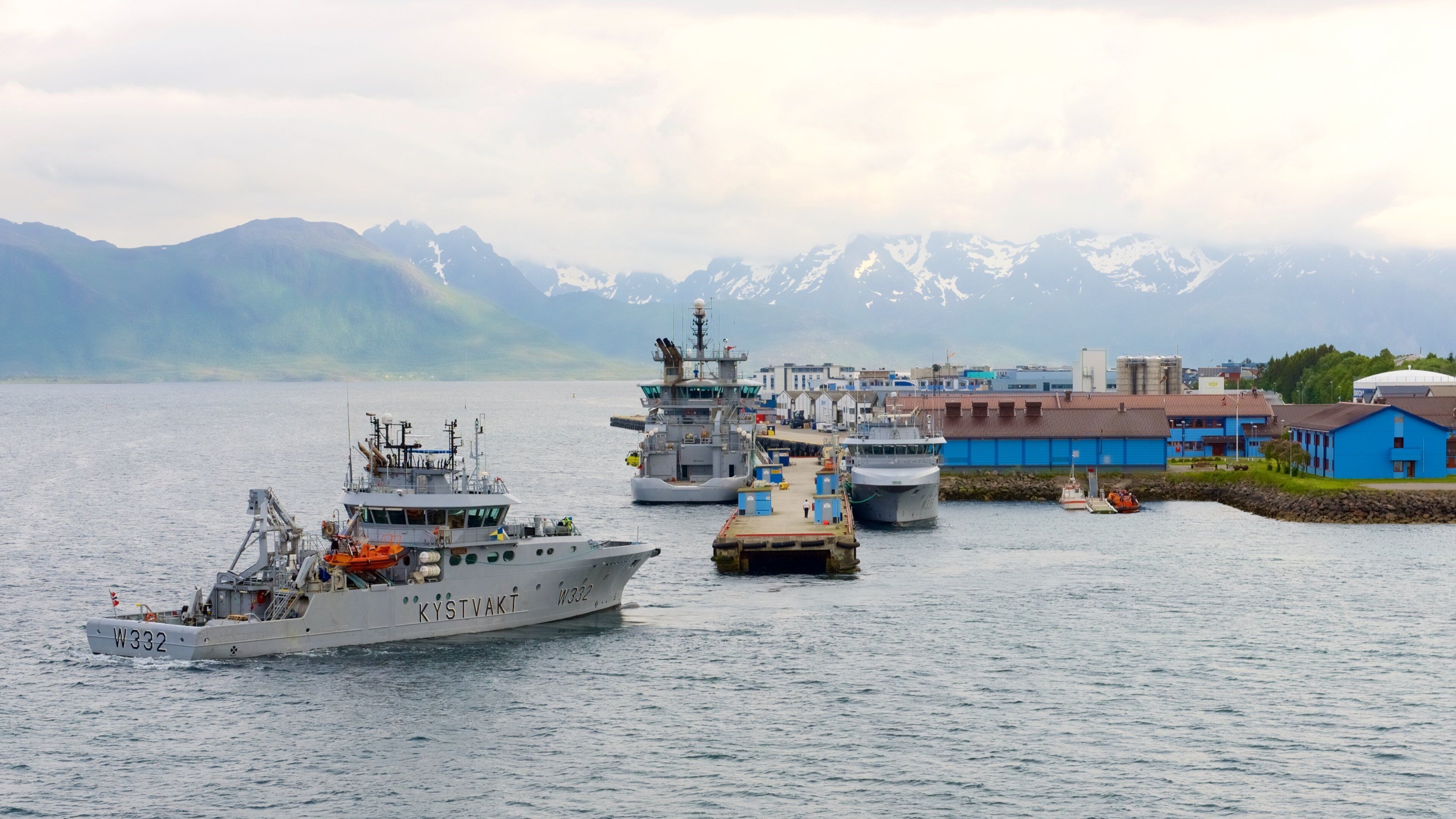 A harbor with several ships docked, a blue building, and snow-capped mountains in the background.