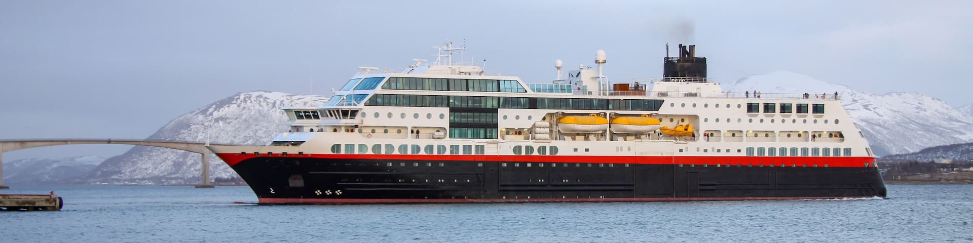 A large cruise ship with a red and black hull, white superstructure, and yellow lifeboats, docked near a bridge and snow-covered mountains.