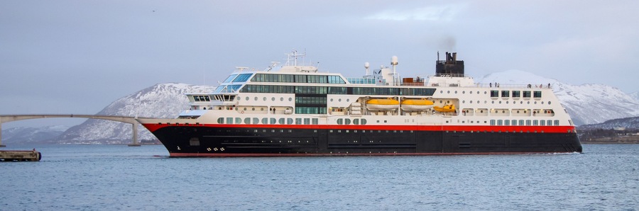 Ein großes Kreuzfahrtschiff mit rotem und schwarzem Rumpf, weißer Aufbauten und gelben Rettungsbooten, liegt an einem Kai in der Nähe einer Brücke und schneebedeckter Berge.
