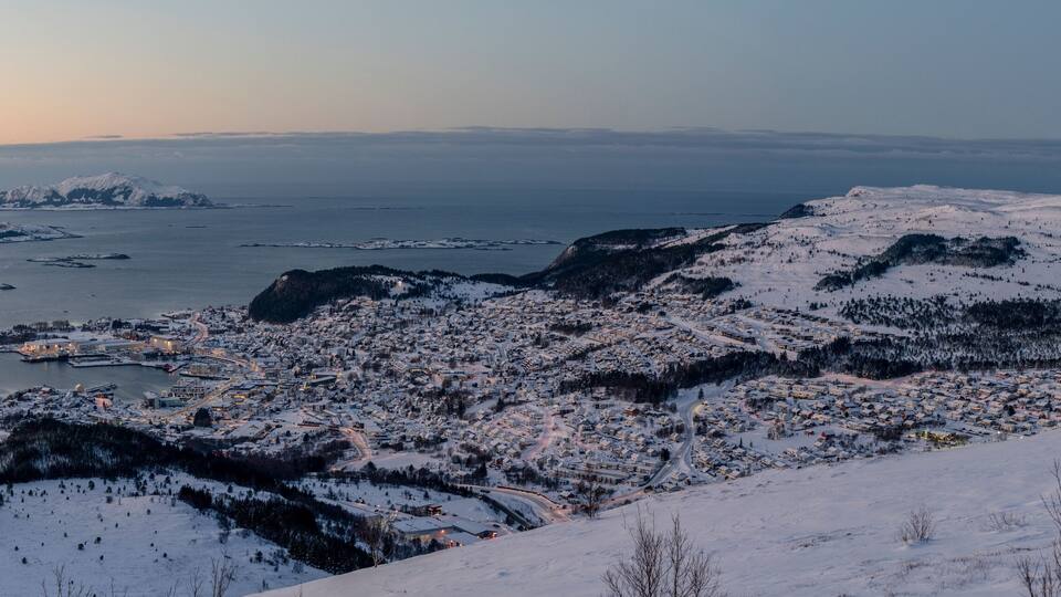 A snow-covered town with a fjord and mountains in the background.