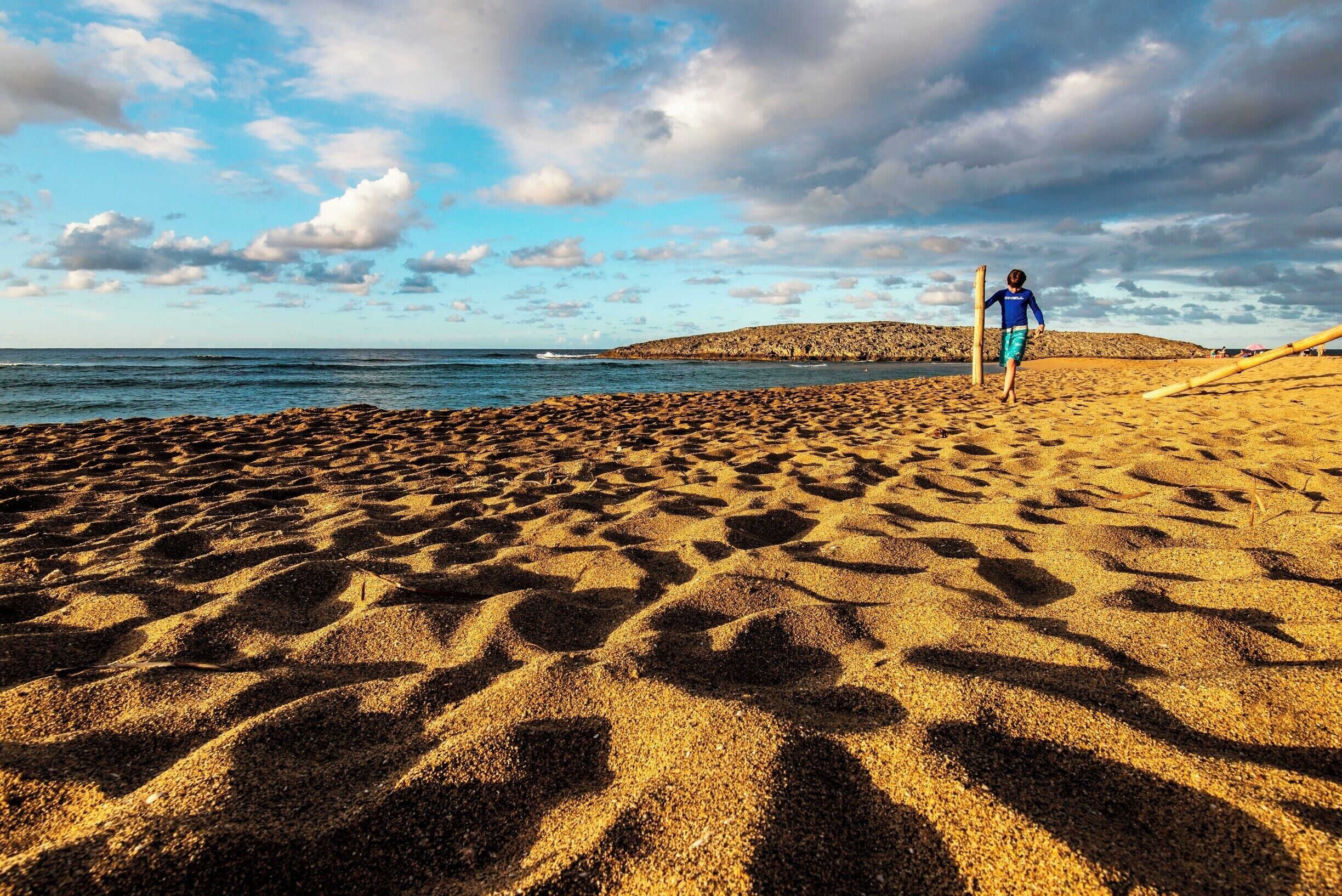 A sandy beach with footprints, a person in blue, and a distant hill.