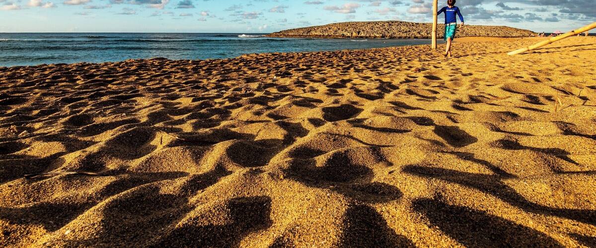 A sandy beach with footprints, a person in blue, and a distant hill.