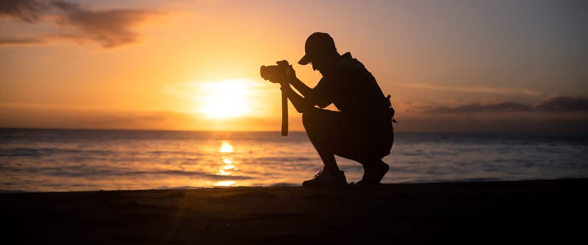 A silhouette of a person taking a photo at sunset by the ocean.