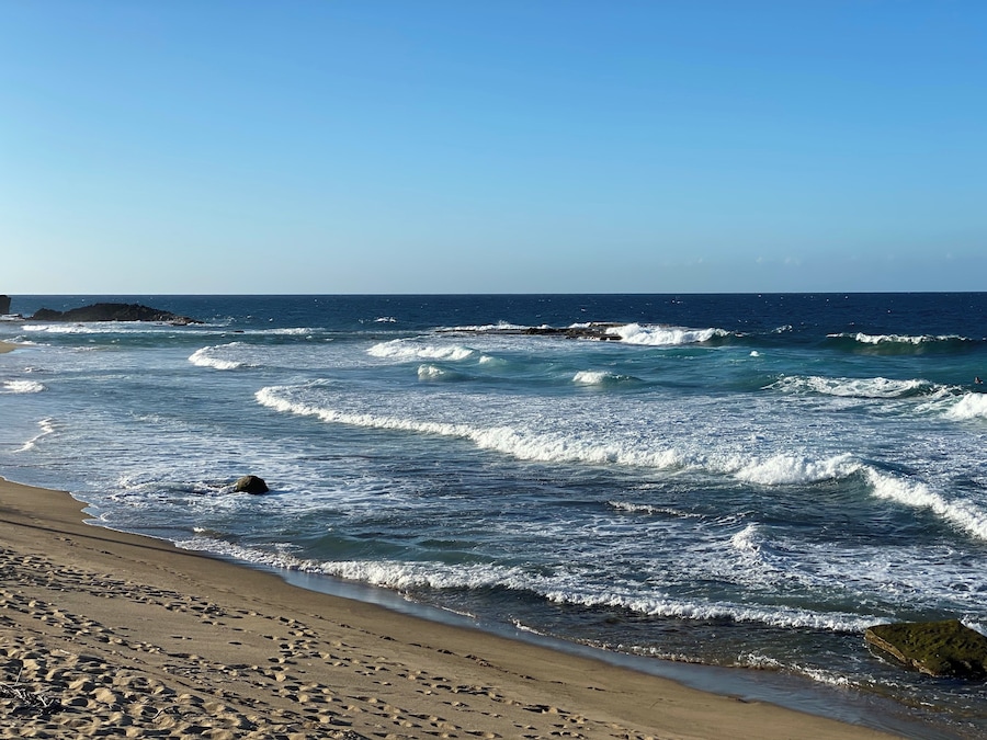 Una playa con olas rompiendo en la orilla, un cielo despejado y afloramientos rocosos a lo lejos.
