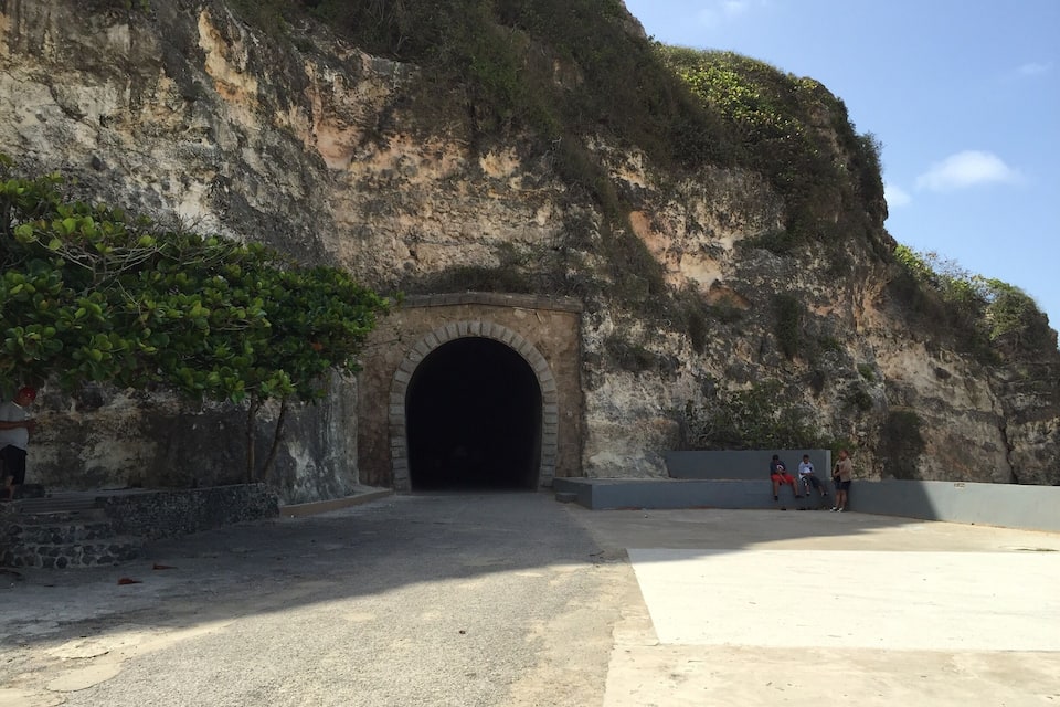 A large cave entrance with a stone archway, surrounded by rocky cliffs and sparse vegetation.