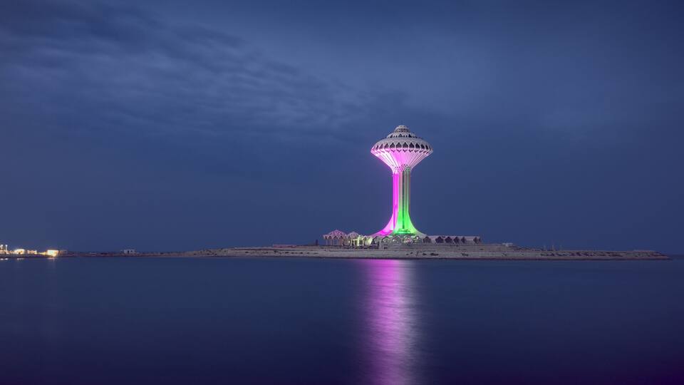 A tall tower with a unique design, illuminated in pink and green lights, standing on a small island in the water.