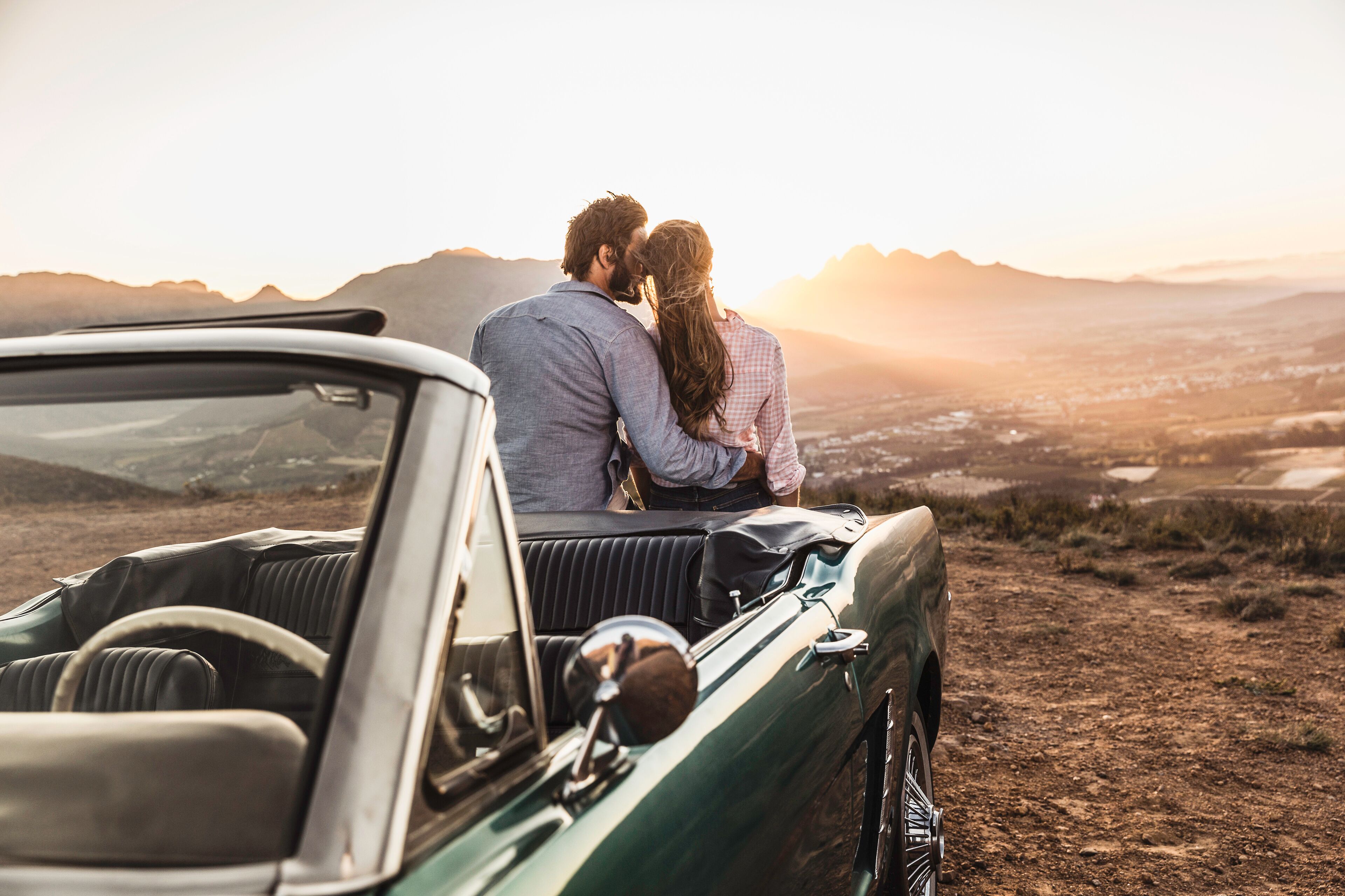 Um casal em um carro conversível apreciando a paisagem montanhosa ao pôr do sol.