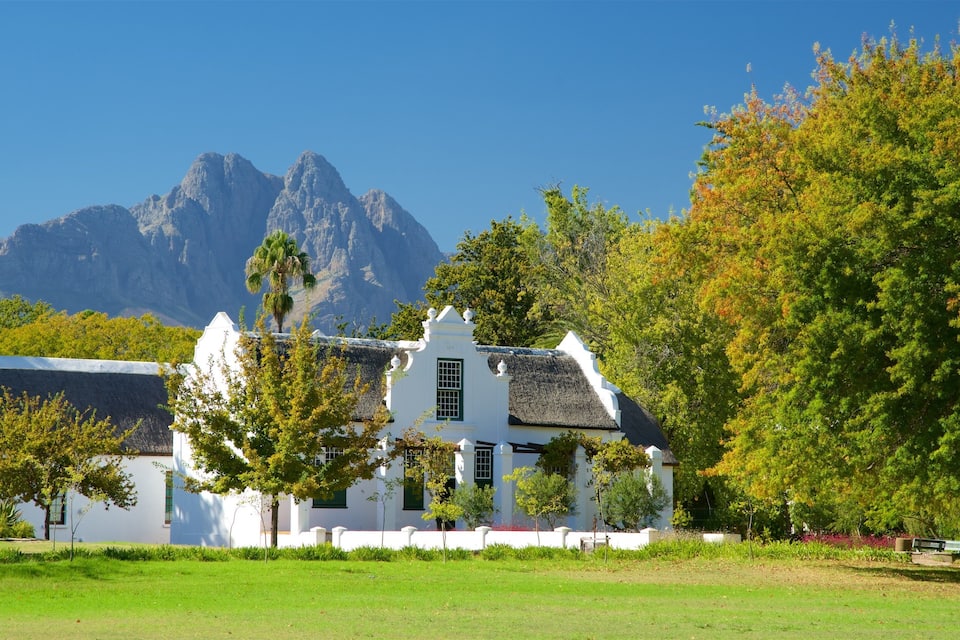 Un edificio blanco con techo de paja, rodeado de árboles y hierba, con montañas al fondo.