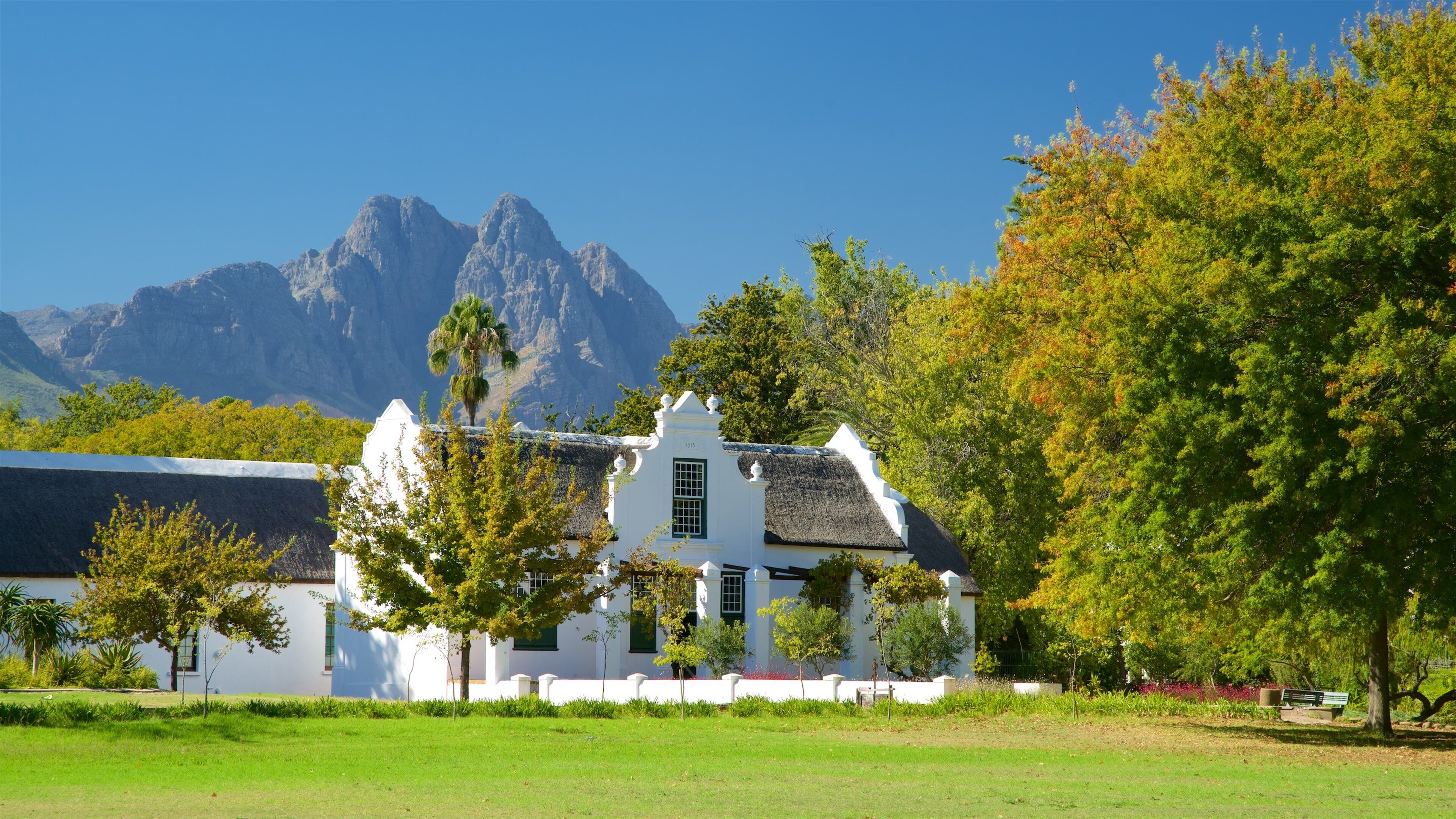 A white building with a thatched roof, surrounded by trees and grass, with mountains in the background.