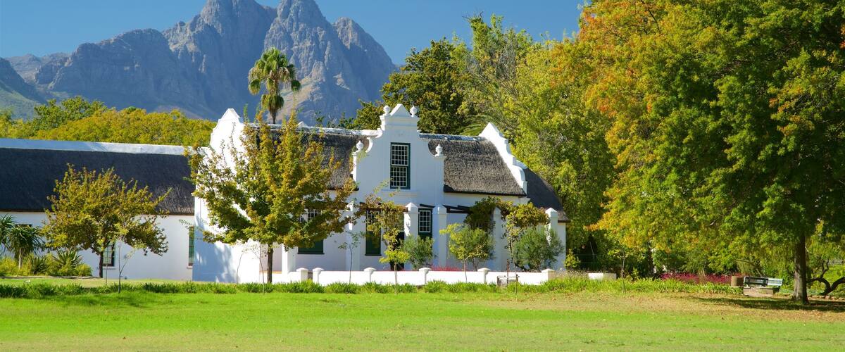 A white building with a thatched roof, surrounded by trees and grass, with mountains in the background.