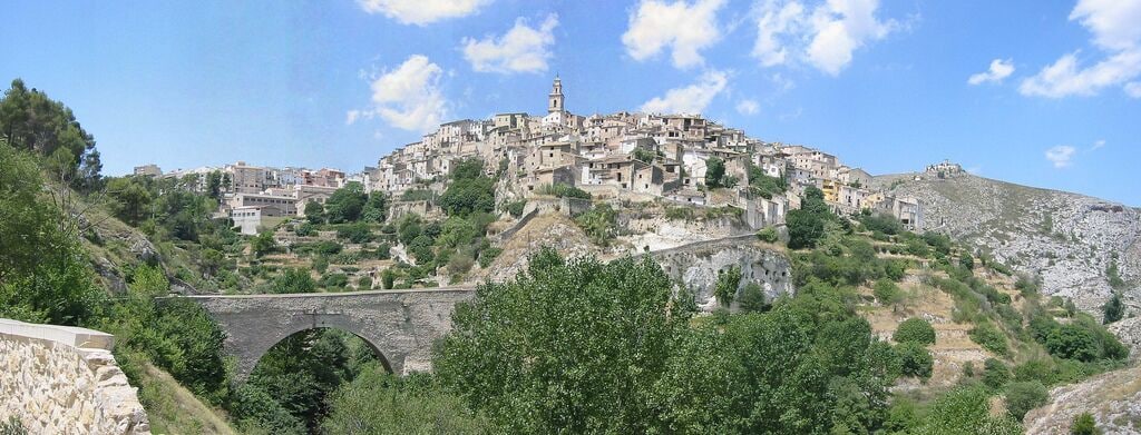 A hillside town with a prominent church on top, a stone bridge, and lush greenery.