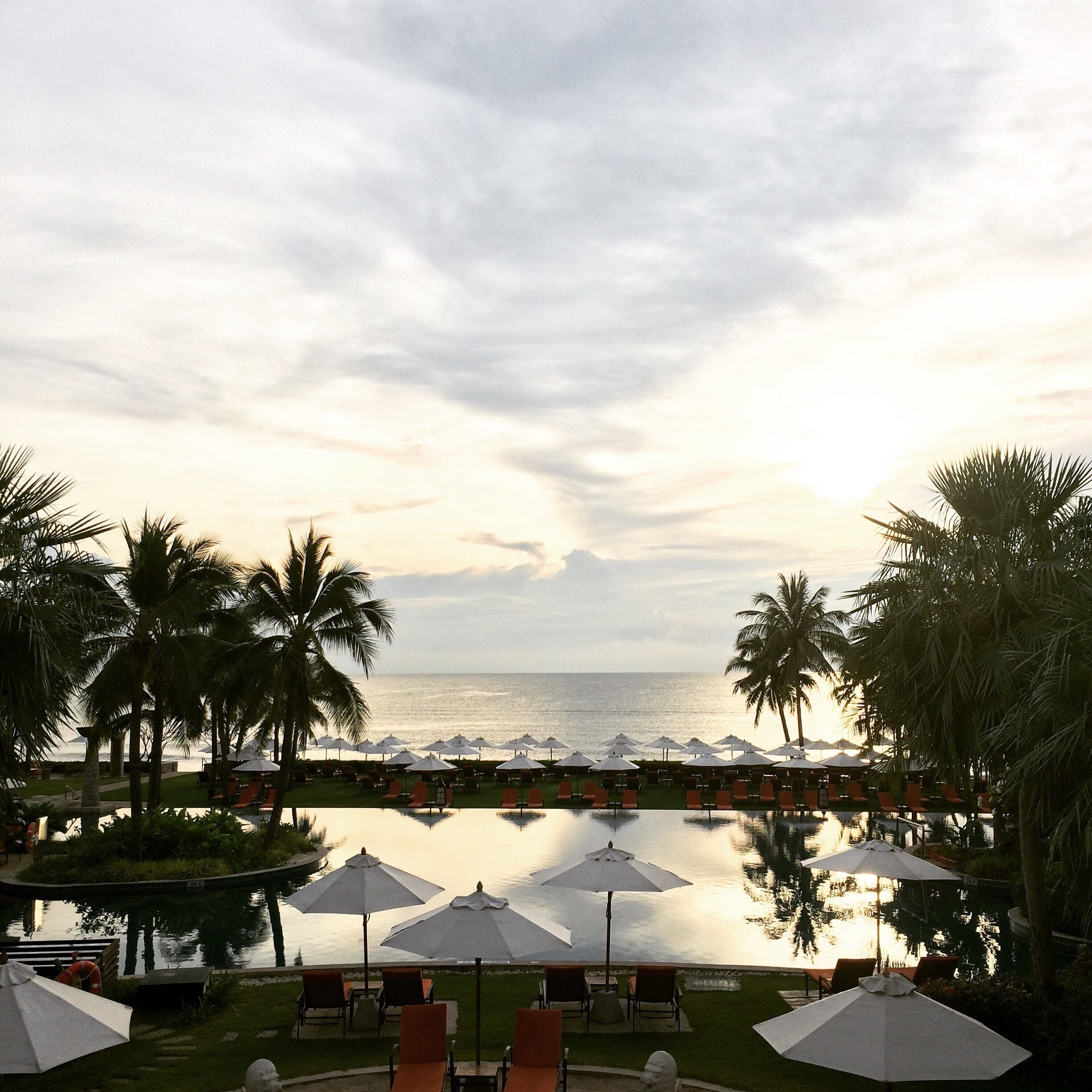 Piscina de un resort con sombrillas y sillas, palmeras y vista al océano.