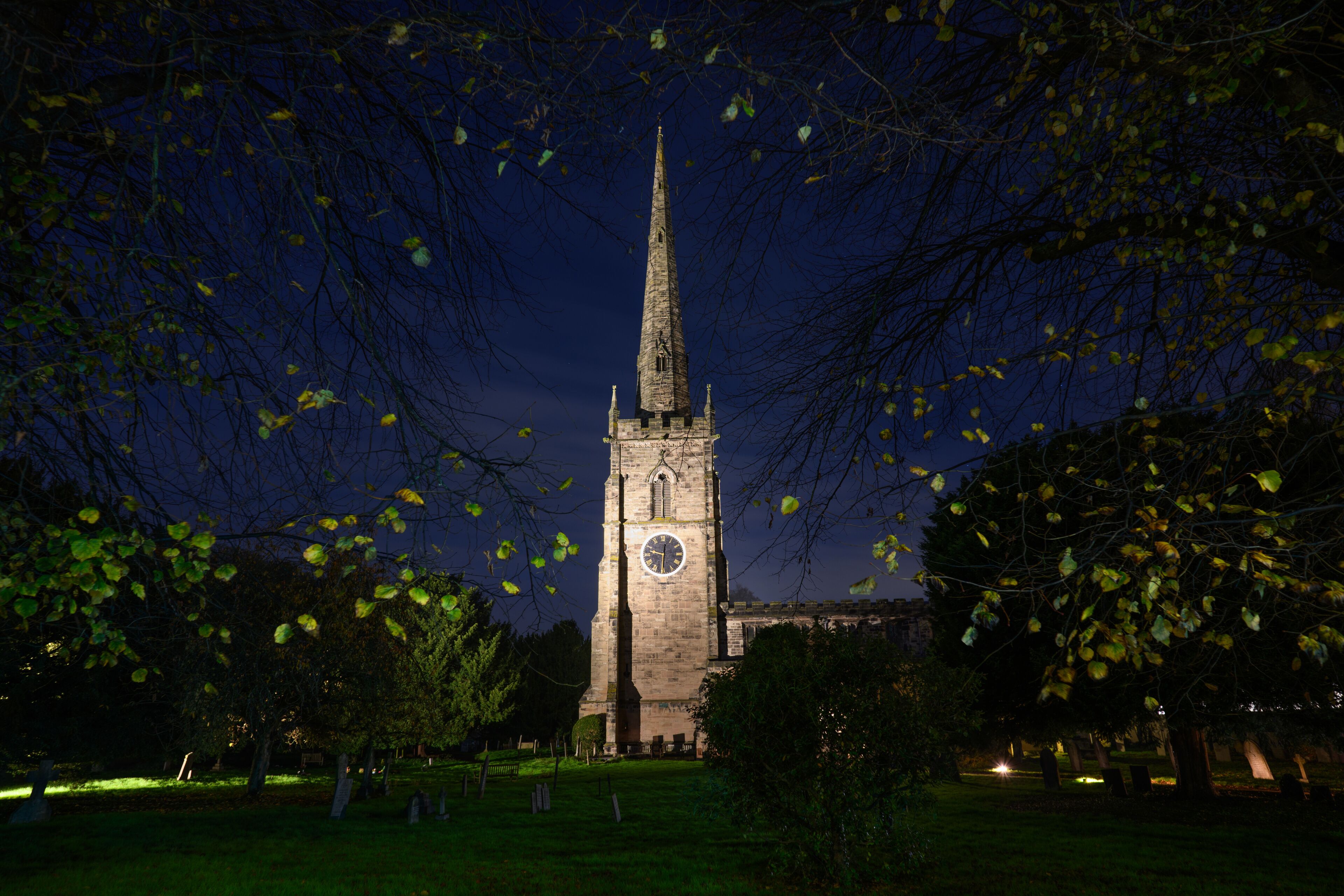 A church tower at night with a clock, surrounded by trees and a cemetery.