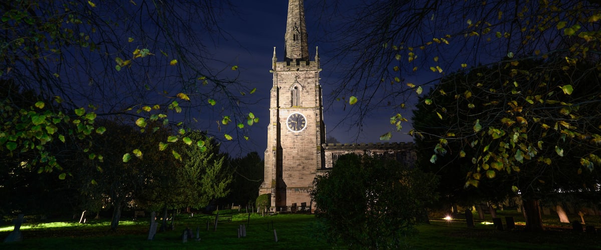 A church tower at night with a clock, surrounded by trees and a cemetery.