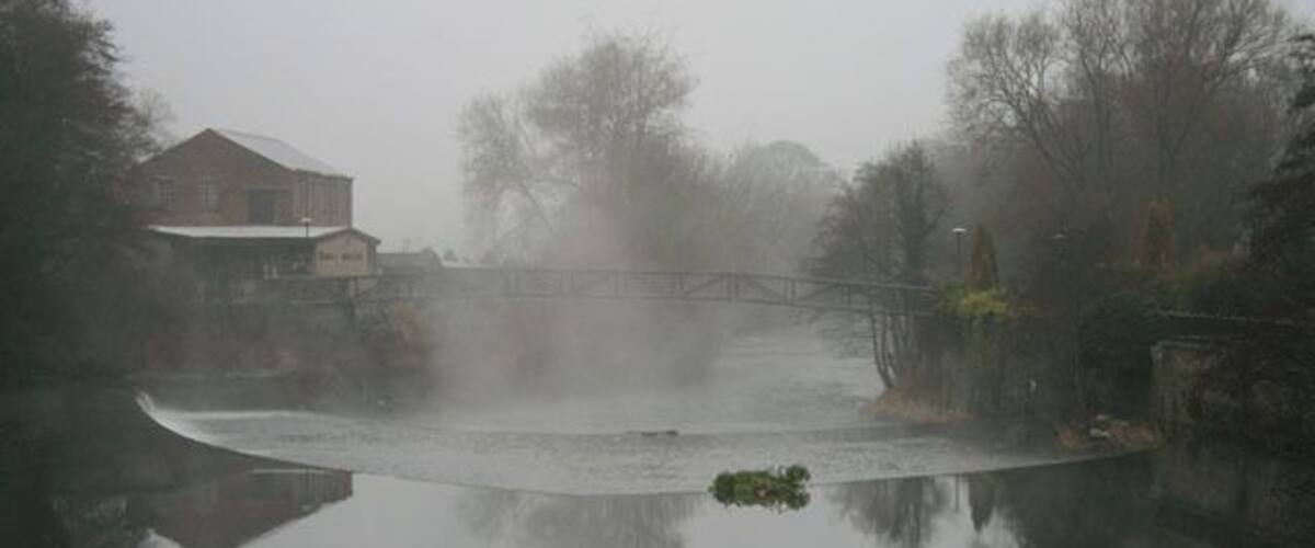 A foggy river with a bridge and a house on the left.