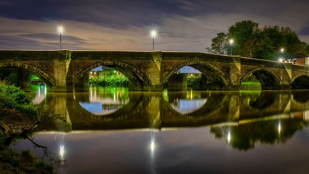 Un pont de pierre à arches enjambant une rivière paisible, dont l’image et les lumières se reflètent dans l’eau.