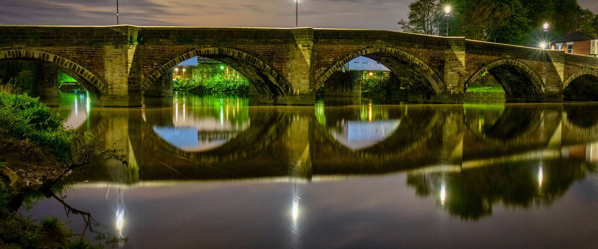 A stone bridge with arches over a calm river, reflecting the bridge and lights.