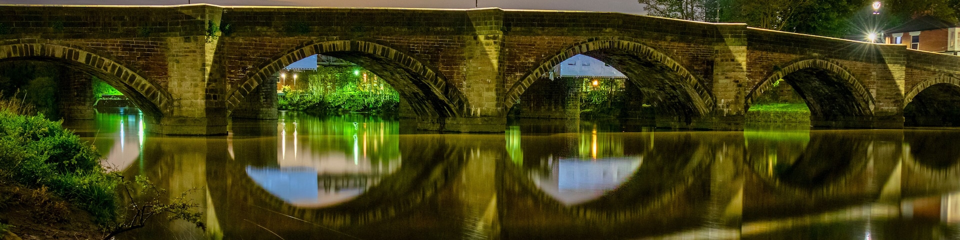 A stone bridge with arches over a calm river, reflecting the bridge and lights.