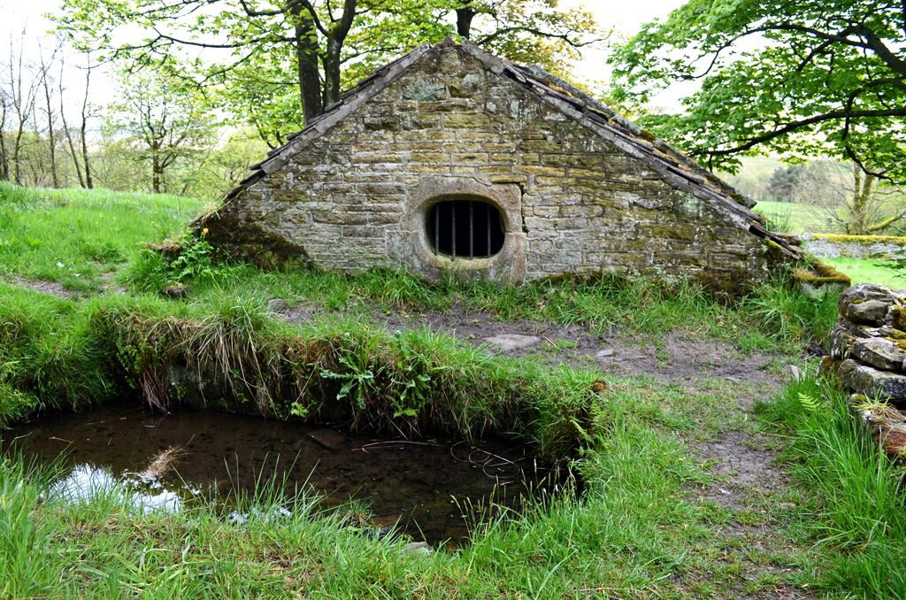 A stone hut with a circular window, surrounded by lush greenery and a small stream.