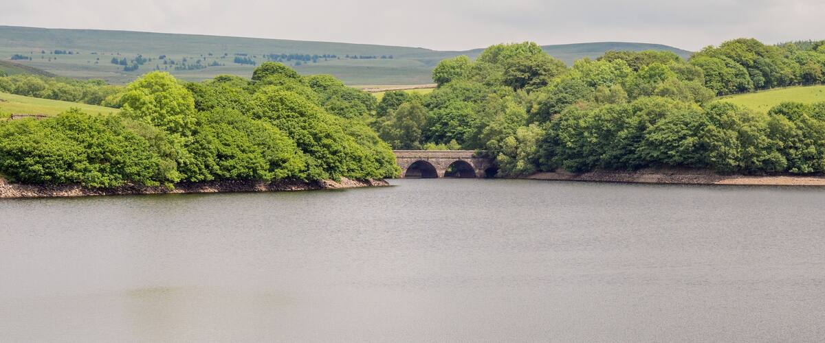 A stone bridge over a calm river, surrounded by lush green hills and dense trees.