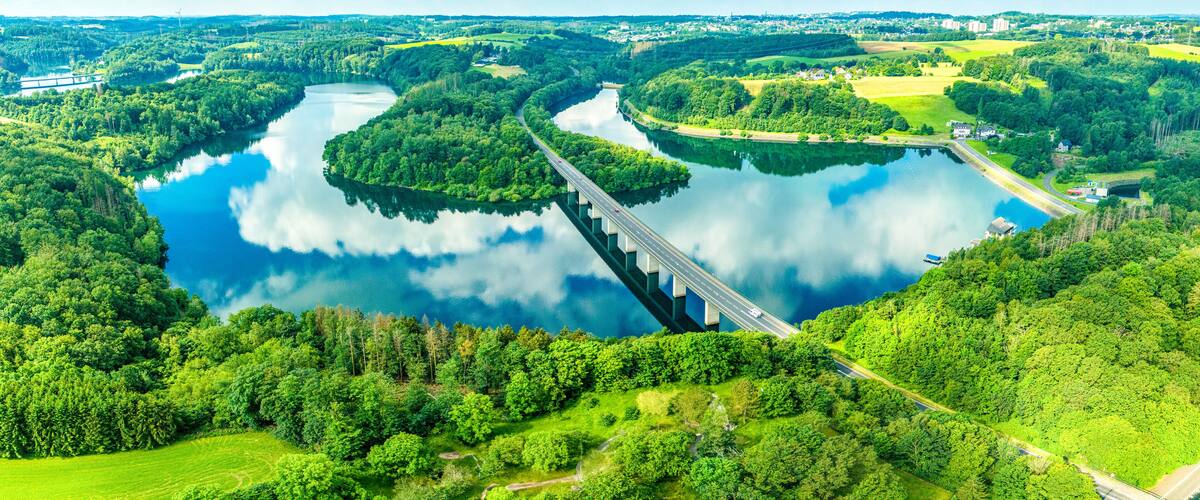 A river with a bridge, surrounded by lush greenery and a cloudy sky.