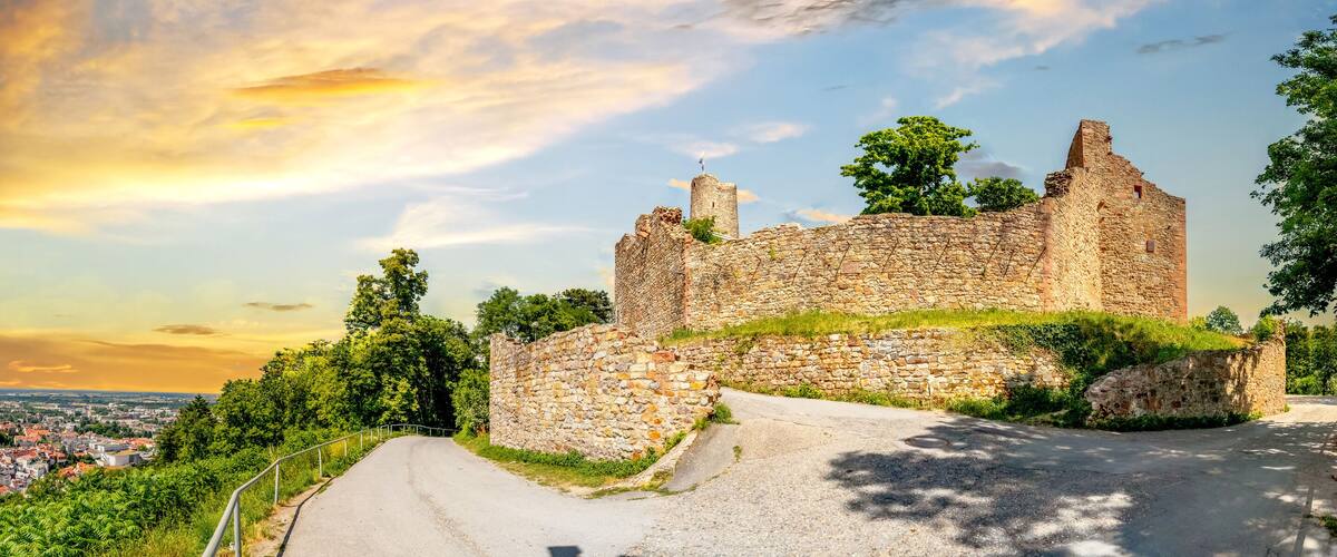 A historic stone castle with a surrounding wall, a paved road leading to it, and a view of a city in the background.