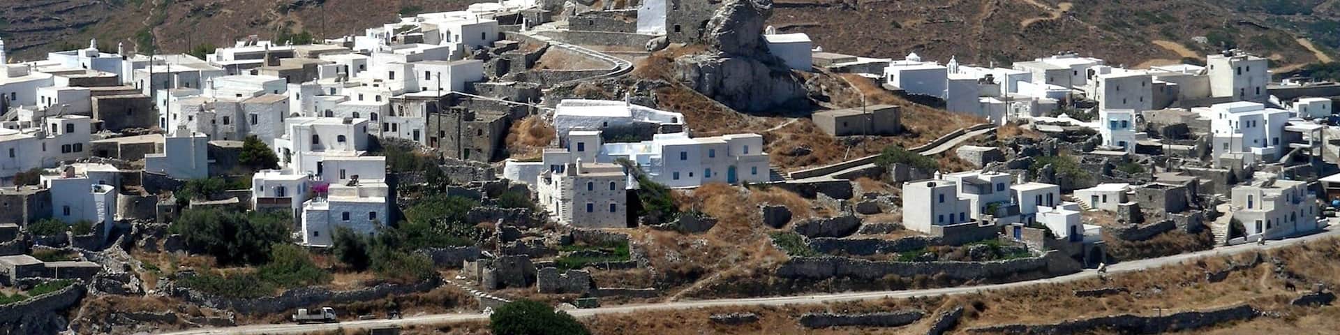 A white-washed village with stone houses nestled on a hillside, overlooking a rugged mountain range and the sea.