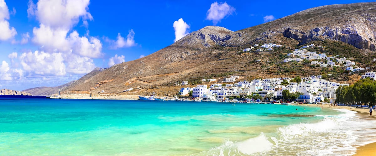 A coastal town with white buildings, a clear blue sea, and a mountainous backdrop.