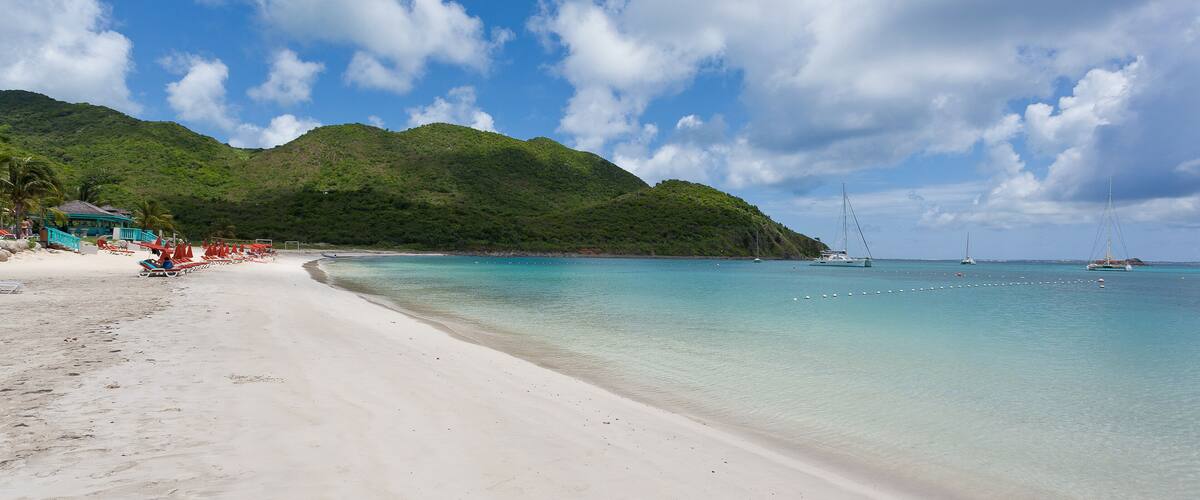 A beach with clear turquoise water, white sand, and green hills in the background.