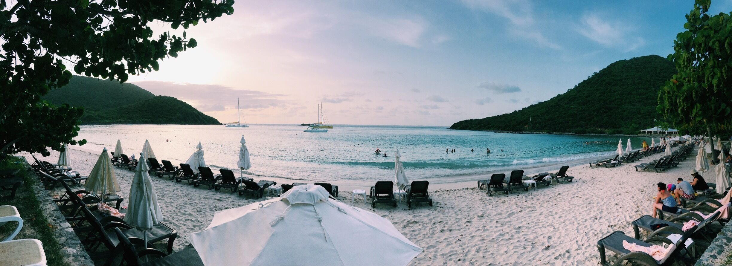 A beach with lounge chairs and umbrellas, boats on the water, and hills in the background.