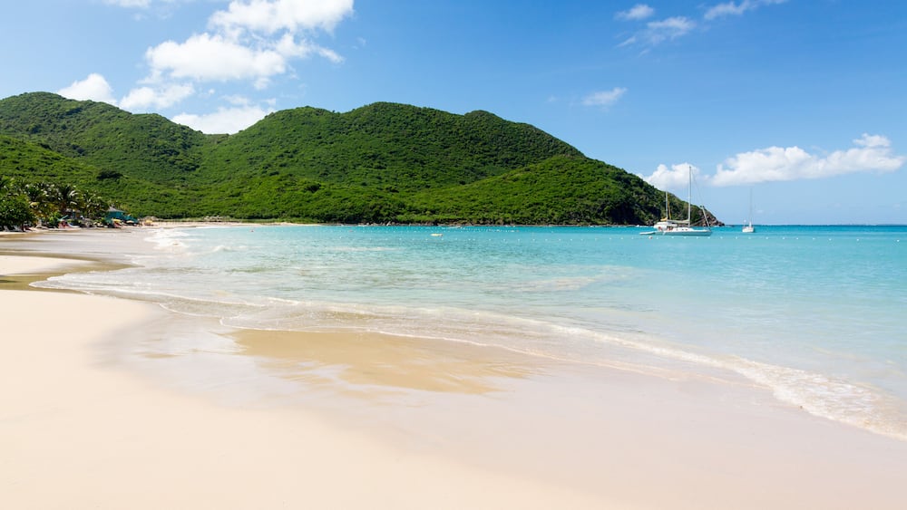 Une plage à l’eau turquoise et limpide, un rivage sablonneux et, en arrière-plan, des collines verdoyantes.