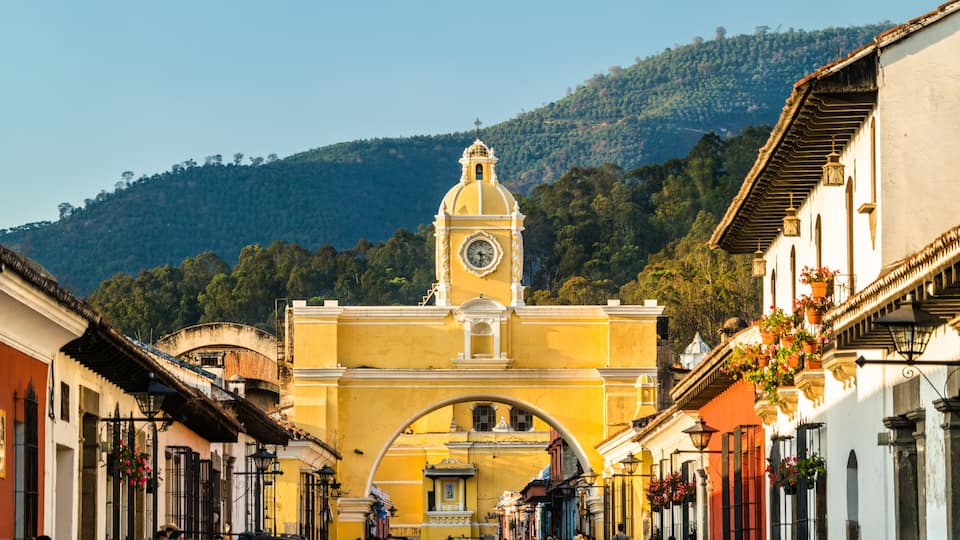 A historic town with a prominent clock tower, a central archway, and a mountainous backdrop.