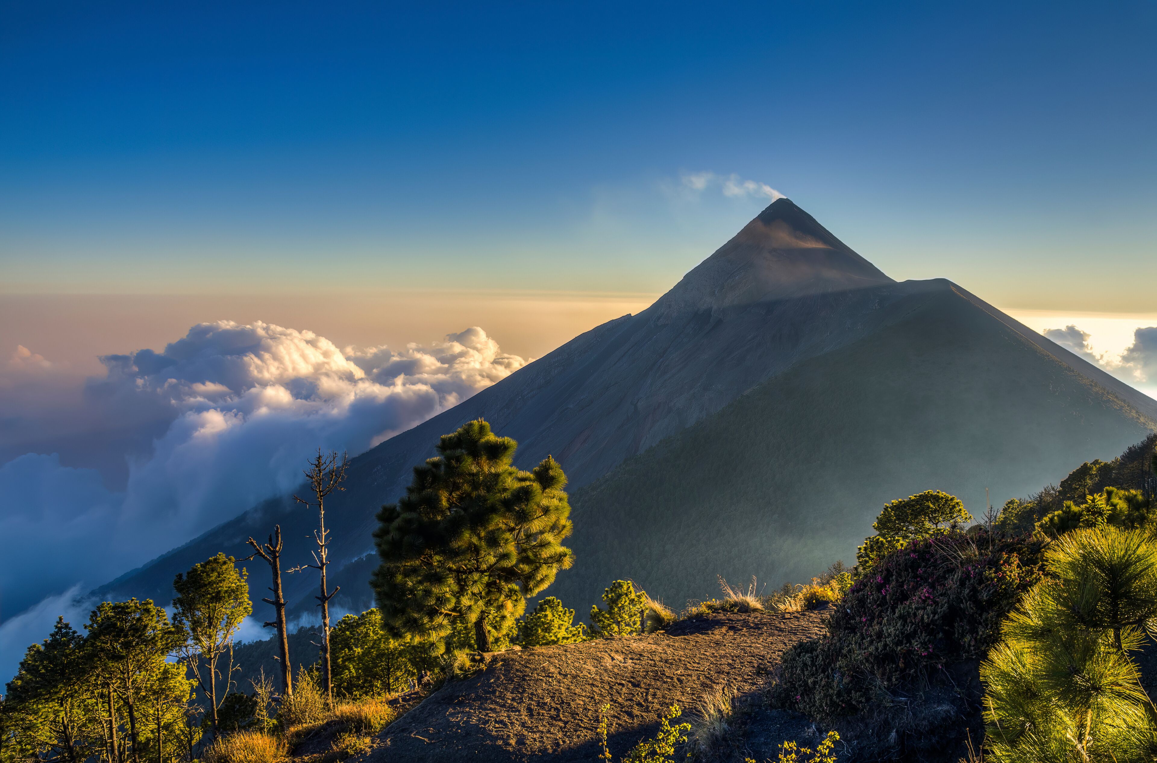 A mountain with a peak, surrounded by clouds and trees.